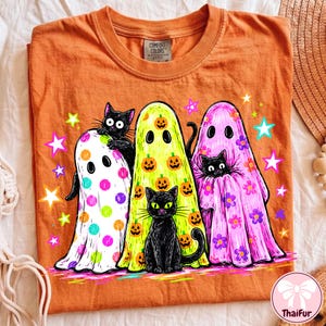 Pu&ograve; includere: Una t-shirt arancione con una grafica raffigurante tre fantasmi e due gatti neri. I fantasmi sono bianchi a pois colorati, gialli con zucche arancioni e rosa con fiori viola. Stelle dai colori vivaci circondano le figure stampate.