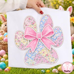 May include: A white fabric item featuring a paw print design with floral patterns in pastel colors. A large pink bow with pearl accents is centered on the paw print. The design is held up by a hand.