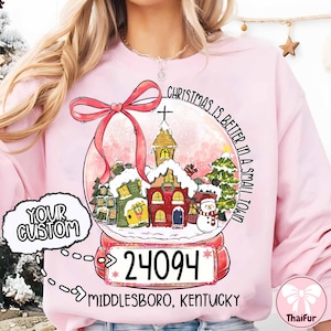 Op de afbeelding: Roze sweatshirt met een sneeuwbolontwerp. De sneeuwbol toont een kleine stad met huizen, een kerk en een sneeuwpop. Tekst luidt "Christmas is better in a small town" en "Middlesboro, Kentucky 24094".