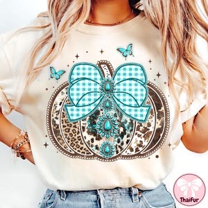 May include: Cream-colored sweatshirt featuring a pumpkin design with a turquoise gingham bow and turquoise gemstone accents. The pumpkin has a leopard print pattern. Three turquoise butterflies and star accents complete the design.