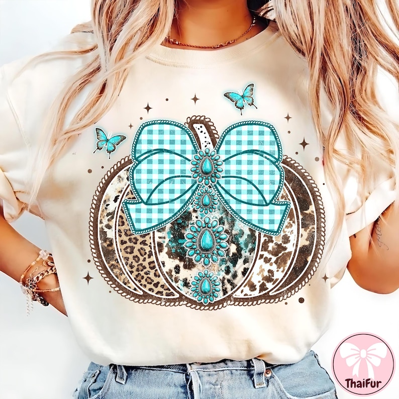 May include: Cream-colored sweatshirt featuring a pumpkin design with a turquoise gingham bow and turquoise gemstone accents. The pumpkin has a leopard print pattern. Three turquoise butterflies and star accents complete the design.