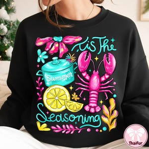 May include: Black sweatshirt with a colorful graphic design. The design includes a pink lobster, a turquoise pot with the word "Summer", a lemon slice, and the text "'Tis The Seasoning".