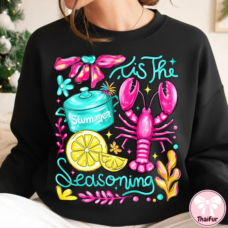 May include: Black sweatshirt with a colorful graphic design. The design includes a pink lobster, a turquoise pot with the word "Summer", a lemon slice, and the text "'Tis The Seasoning".