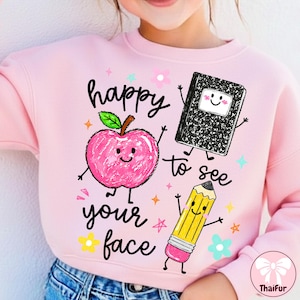May include: A light pink sweatshirt featuring a graphic print of a smiling pink apple, a black and white composition notebook, and a yellow pencil. The text "happy to see your face" is printed in black script, surrounded by small colorful stars and flowers.