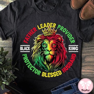May include: Dark gray t-shirt featuring a lion's head with a crown, in red, yellow, and green. Text around the lion reads "Father, Leader, Provider, Protector, Blessed, Husband, King, Black."