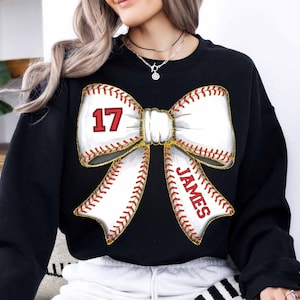 May include: Black sweatshirt with a white baseball-themed bow. The bow has red stitching and the number "17" on one side and the name "JAMES" on the other.