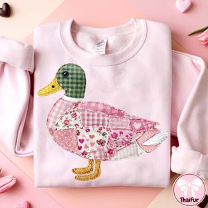 Può includere: Una felpa rosa chiaro con un motivo a patchwork di anatra. L'anatra ha una testa a scacchi verdi, un becco giallo e un corpo fatto di vari tessuti rosa a fantasia. La felpa ha maniche lunghe e girocollo.