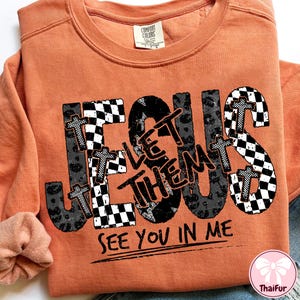 May include: Orange crewneck sweatshirt with the word "JESUS" in a mix of checkered, leopard print, and cross designs. The text "LET THEM SEE YOU IN ME" is printed below the word JESUS.