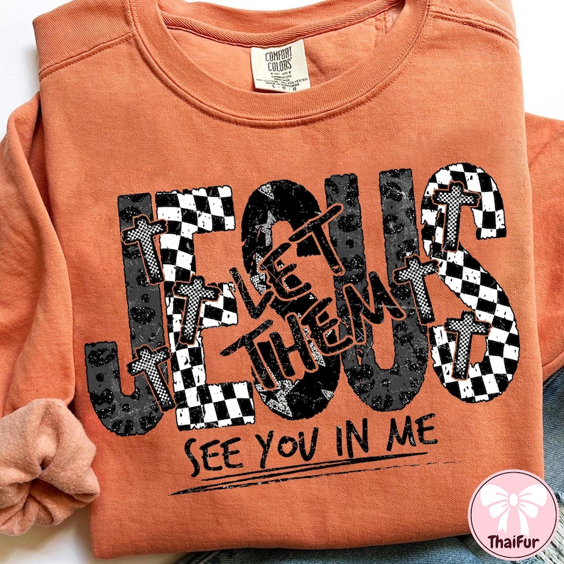 May include: Orange crewneck sweatshirt with the word "JESUS" in a mix of checkered, leopard print, and cross designs. The text "LET THEM SEE YOU IN ME" is printed below the word JESUS.