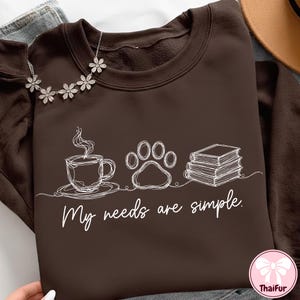 May include: A brown sweatshirt with a white line drawing of a coffee cup, paw print, and books. The text "My needs are simple" is below the drawings. A silver flower necklace is draped on the sweatshirt.