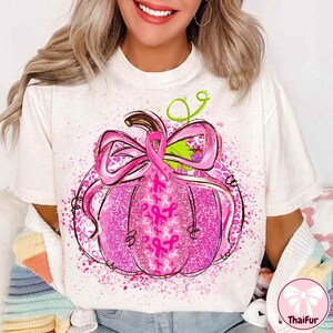 May include: White t-shirt featuring a pink pumpkin design with a large bow and pink ribbons. The pumpkin has a glittery texture and is decorated with pink ribbons, symbolizing breast cancer awareness. The design includes green vines and a brown stem.