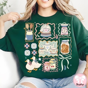 Peut inclure: Sweat-shirt vert fonc&eacute; avec un motif sur le th&egrave;me de la cuisine. Le motif comprend des illustrations d'ustensiles de cuisine, de tasses &agrave; th&eacute;, de biscuits et de canards. L'esth&eacute;tique g&eacute;n&eacute;rale est de style vintage.