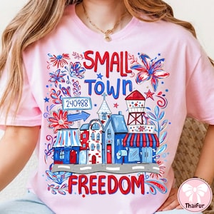 May include: Light pink t-shirt with a colorful graphic design. The design features the words "SMALL TOWN FREEDOM" with illustrations of buildings, flowers, and stars in red, white, and blue. A whimsical and patriotic theme.