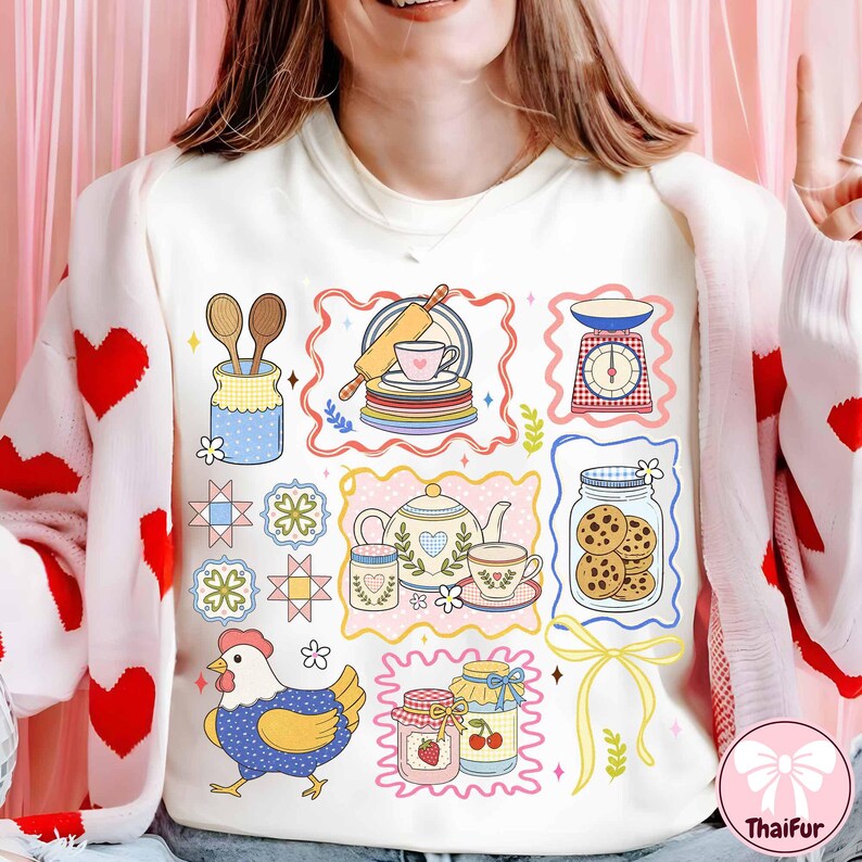 May include: Cream sweatshirt with a colourful, illustrated design of kitchen-themed elements. The design includes a chicken, jars of cookies and jam, a teapot, and a scale. The design is framed with decorative borders.