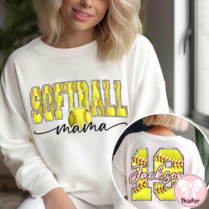 Op de afbeelding: Cr&egrave;mekleurige sweatshirt met de woorden "SOFTBALL mama" in gele en zwarte letters, met een softbalafbeelding. De achterkant van de sweatshirt heeft het nummer 18 en de naam "Jackson" in een vergelijkbare stijl.