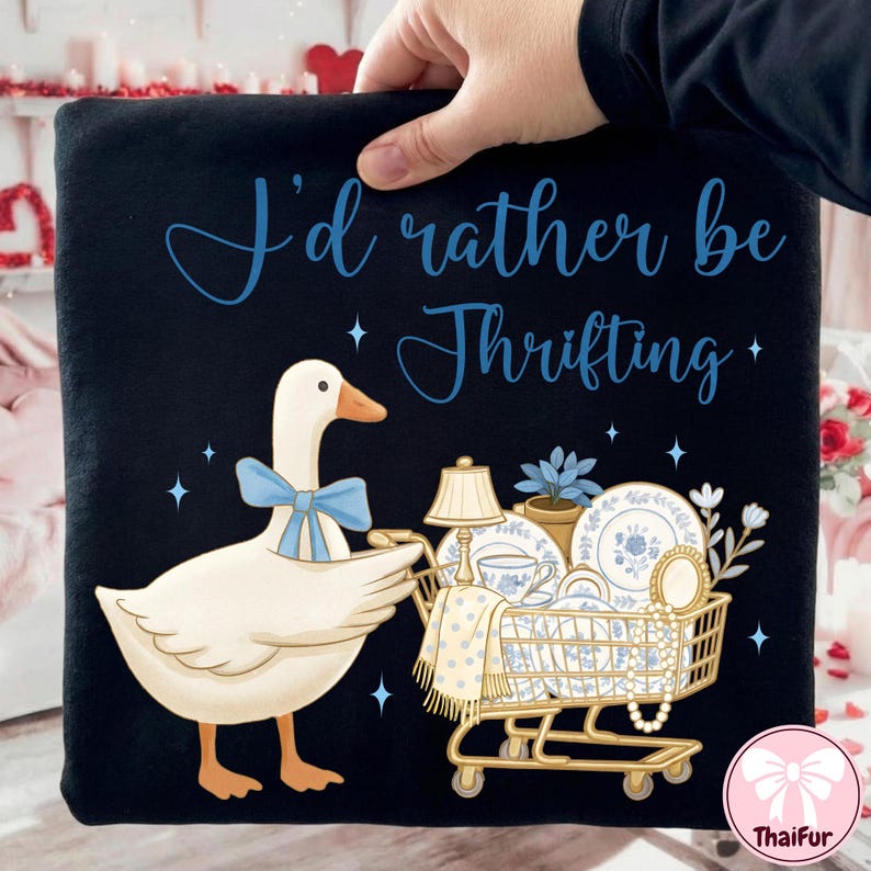 May include: Black graphic tee featuring a cartoon goose with a blue bow tie pushing a shopping cart filled with vintage items. The text "I'd rather be Thrifting" is in blue script above the goose.