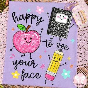 May include: A light purple t-shirt featuring a graphic print with the text "happy to see your face" in black script. The design includes a pink apple, a black and white composition notebook, and a yellow pencil, all with smiling faces.