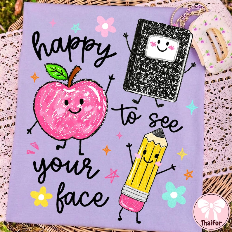 May include: A light purple t-shirt featuring a graphic print with the text "happy to see your face" in black script. The design includes a pink apple, a black and white composition notebook, and a yellow pencil, all with smiling faces.