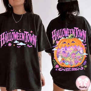 May include: A black t-shirt featuring the text HALLOWEENTOWN in pink on the front. The back displays a large orange jack-o'-lantern graphic containing a colorful town scene with ghosts, a scarecrow, and the text HALLOWEENTOWN EST. 1998 in pink.