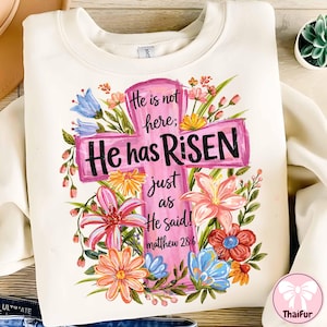 Puede incluir: Sudadera color crema con un diseño de cruz rosa con las palabras "He has Risen" y detalles florales. También incluye el texto "He is not here; just as He said! Matthew 28:6". El diseño incorpora varias flores coloridas.