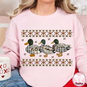 May include: A light pink sweatshirt featuring three ducks with plaid patterns and green heads. The ducks are arranged in a row, with a decorative border of hearts and squares above and below them. The sweatshirt has long sleeves.