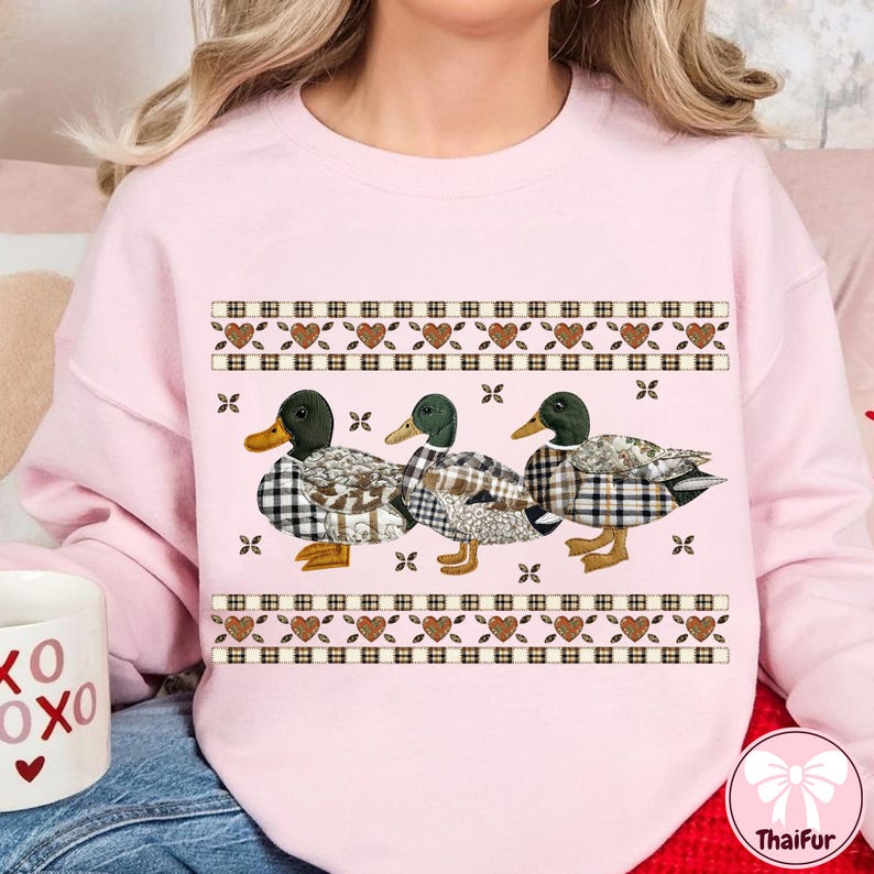 May include: A light pink sweatshirt featuring three ducks with plaid patterns and green heads. The ducks are arranged in a row, with a decorative border of hearts and squares above and below them. The sweatshirt has long sleeves.
