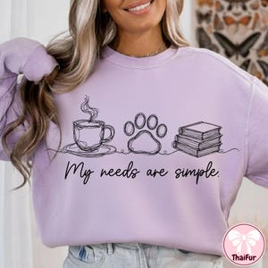 May include: A light purple sweatshirt with a black line drawing of a coffee cup, paw print, and books. The text "My needs are simple" is below the drawings. The sweatshirt is a casual style.