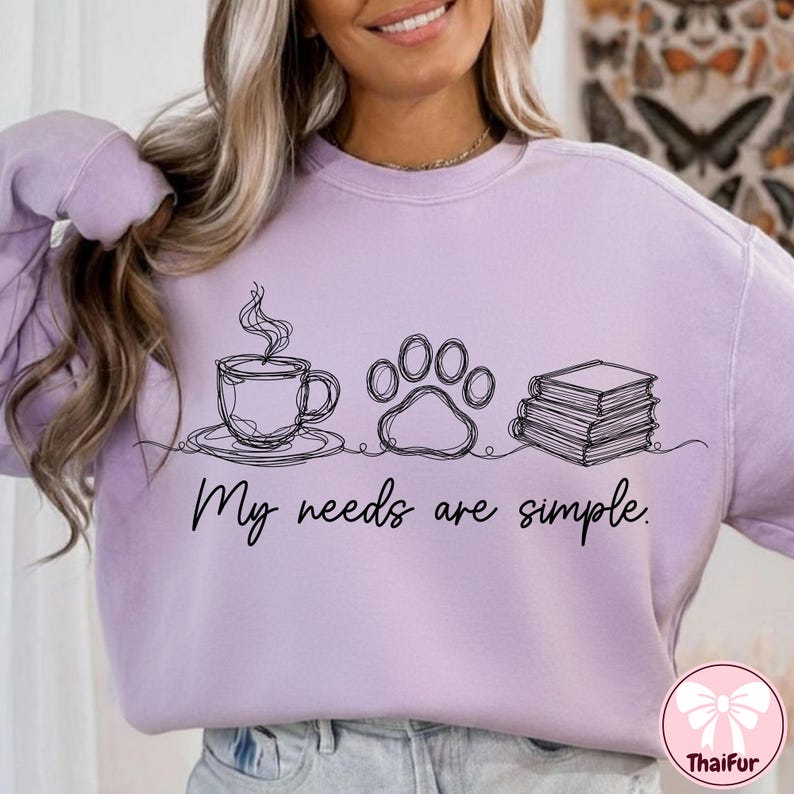 May include: A light purple sweatshirt with a black line drawing of a coffee cup, paw print, and books. The text "My needs are simple" is below the drawings. The sweatshirt is a casual style.