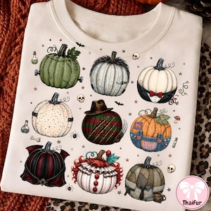 May include: A cream-colored t-shirt featuring a graphic print of nine stylized pumpkins inspired by horror movie characters. The design includes various pumpkin illustrations with details like stitches, hats, overalls, and suits, surrounded by small stars, skulls, and potion bottles.