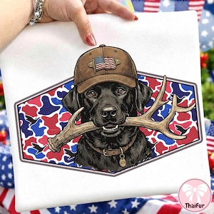 Può includere: T-shirt bianco con grafica di un Labrador Retriever nero che indossa un berretto marrone con un disegno della bandiera americana. Il cane tiene le corna in bocca, su uno sfondo mimetico rosso, bianco e blu. La maglietta ha un colletto marrone.