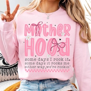 May include: Pink sweatshirt with the text "Mother Hood" in pink letters with black outlines and a pink bow. The text "some days I rock it, some days it rocks me, either way we're rockin'" is printed below the main text.