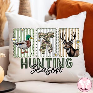 Puede incluir: Coj&iacute;n blanco con fondo a rayas verdes y blancas. Presenta tres im&aacute;genes enmarcadas: un pato, un lazo de camuflaje y un ciervo. Las palabras "HUNTING Season" est&aacute;n impresas en verde y negro.