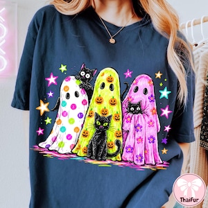 Pu&ograve; includere: Una t-shirt blu scuro con una stampa grafica di tre fantasmi e tre gatti neri. Un fantasma &egrave; bianco a pois colorati, uno &egrave; giallo con zucche arancioni e uno &egrave; rosa con fiori viola, circondati da stelle colorate.