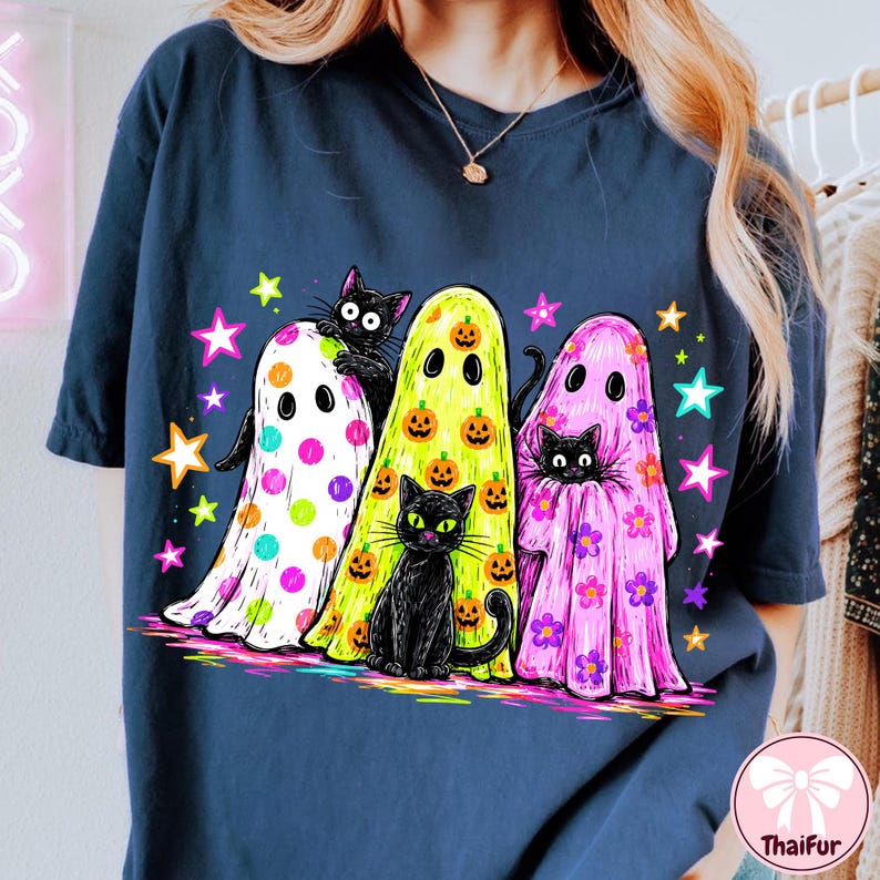 Pu&ograve; includere: Una t-shirt blu scuro con una stampa grafica di tre fantasmi e tre gatti neri. Un fantasma &egrave; bianco a pois colorati, uno &egrave; giallo con zucche arancioni e uno &egrave; rosa con fiori viola, circondati da stelle colorate.