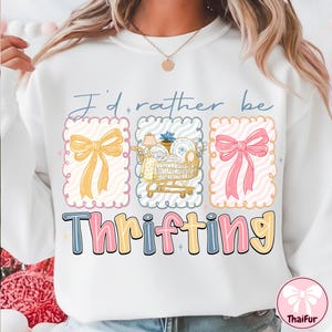 May include: White sweatshirt with the text "I'd rather be thrifting." The design includes three framed illustrations: a yellow bow, a shopping cart filled with items, and a pink bow. The word "Thrifting" is in colorful block letters.