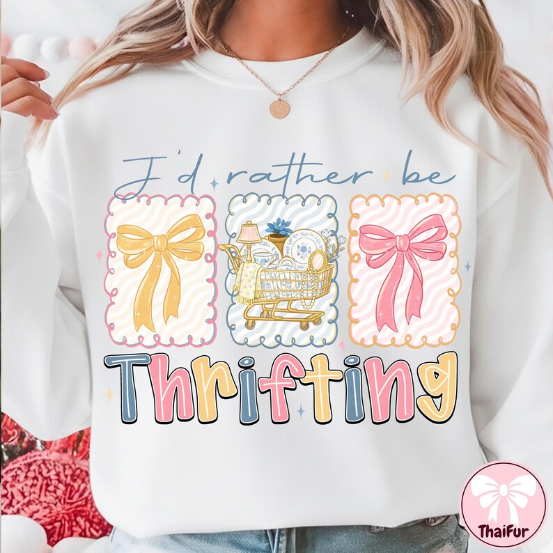 May include: White sweatshirt with the text "I'd rather be thrifting." The design includes three framed illustrations: a yellow bow, a shopping cart filled with items, and a pink bow. The word "Thrifting" is in colorful block letters.