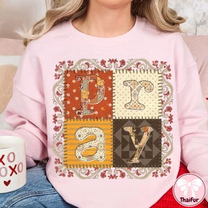 May include: A light pink sweatshirt with the word "PRAY" in a patchwork design. Each letter is in a different patterned square, with a floral border. The sweatshirt has a crew neck.