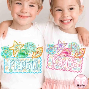 Peut inclure: Deux enfants portant des t-shirts blancs avec un motif de coquillages colorés. Les chemises présentent une étoile de mer, des coquillages et les noms "Martin" et "Kayce" dans une police décorative. Le design comprend des couleurs bleues, vertes, jaunes et roses.