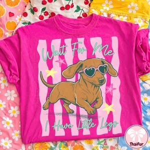 Op de afbeelding: Felroze T-shirt met een bruine teckel met een hartvormige zonnebril. De tekst "Wait For Me, I Have Little Legs" staat erbij, met sterren en strepen op de achtergrond.