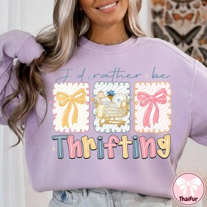 May include: A light purple sweatshirt with the text "I'd rather be thrifting." The design includes three framed illustrations: a yellow bow, a shopping cart with dishes, and a pink bow. The word "Thrifting" is in colorful bubble letters.