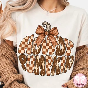 May include: A cream-colored t-shirt featuring a large graphic of a pumpkin shape filled with a brown and white checkered pattern, small cactus icons, and stars. The text HOWDY PUMPKIN is integrated into the design, topped with a brown floral bow.