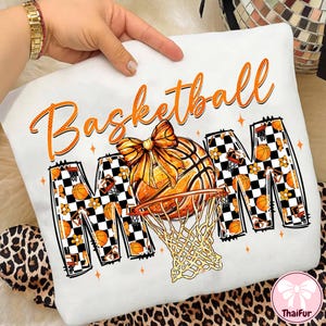May include: White sweatshirt with an orange basketball design. The word "Basketball" is written in orange script above the image of a basketball and hoop. The word "MOM" is in a checkered pattern with basketballs and flowers.