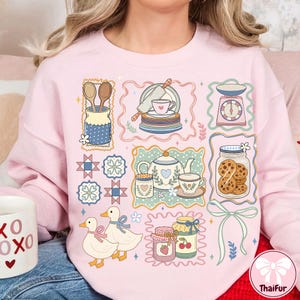 Peut inclure: Un sweat-shirt rose clair orn&eacute; d'un collage d'illustrations sur le th&egrave;me de la cuisine. Le motif comprend des images d'ustensiles de cuisine, d'une th&eacute;i&egrave;re, de biscuits, de canards et de pots de confiture. L'esth&eacute;tique g&eacute;n&eacute;rale est mignonne et fantaisiste.