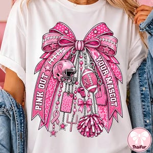 Puede incluir: Una camiseta blanca con un gran gráfico rosa de un lazo con dijes de fútbol americano, incluyendo un casco, un balón, un silbato, un pompón y un poste de meta. El texto PINK OUT YOUR MASCOT aparece en letras rosas sobre las cintas.