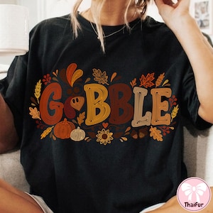 May include: A black short-sleeved t-shirt featuring the word GOBBLE in large, multi-colored, line-art style letters. The design is surrounded by autumn-themed illustrations including maple leaves, wheat stalks, pumpkins, a sunflower, and acorns.