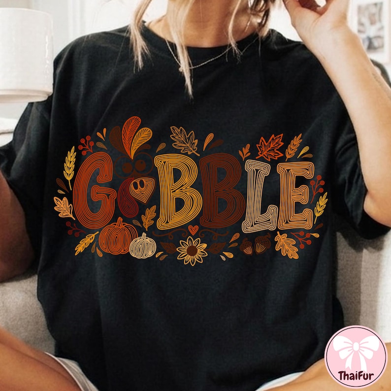 May include: A black short-sleeved t-shirt featuring the word GOBBLE in large, multi-colored, line-art style letters. The design is surrounded by autumn-themed illustrations including maple leaves, wheat stalks, pumpkins, a sunflower, and acorns.