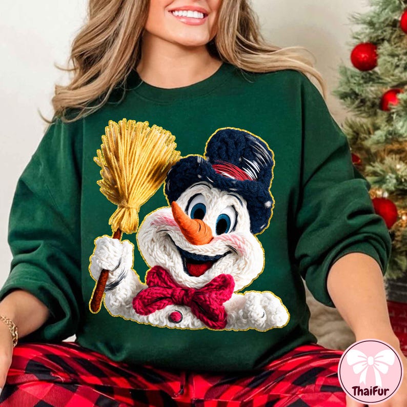 May include: Green sweatshirt with a cheerful snowman design. The snowman has a carrot nose, blue eyes, and a black top hat. It holds a yellow broom and wears a red bow tie. A festive Christmas jumper.