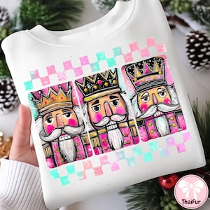 K&ouml;nnte beinhalten: Wei&szlig;es Sweatshirt mit einem farbenfrohen Aufdruck von drei Nussknackersoldaten. Das Design umfasst rosa und goldfarbene Akzente sowie ein rosa, blaues und wei&szlig;es Karomuster. Die Nussknacker haben wei&szlig;e Schnurrb&auml;rte und tragen Kronen.