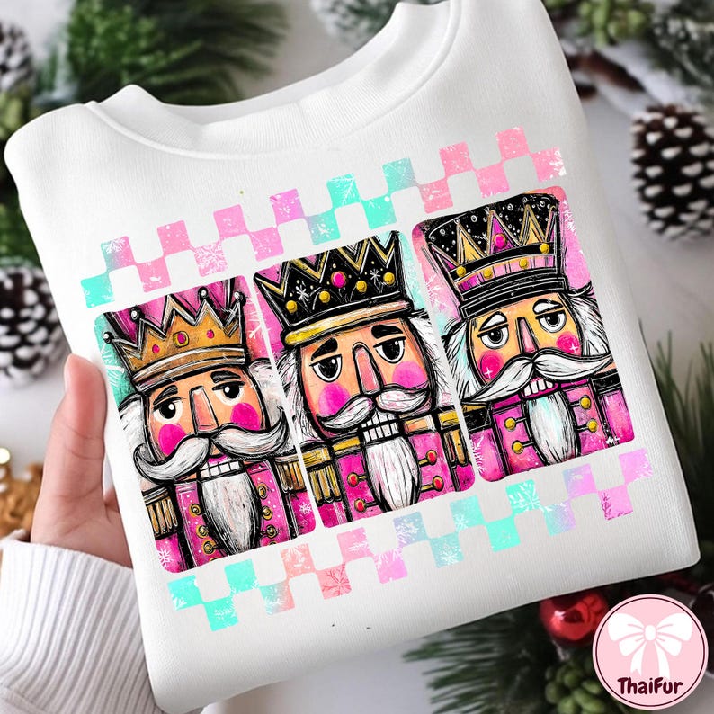 K&ouml;nnte beinhalten: Wei&szlig;es Sweatshirt mit einem farbenfrohen Aufdruck von drei Nussknackersoldaten. Das Design umfasst rosa und goldfarbene Akzente sowie ein rosa, blaues und wei&szlig;es Karomuster. Die Nussknacker haben wei&szlig;e Schnurrb&auml;rte und tragen Kronen.