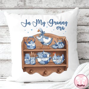K&ouml;nnte beinhalten: Wei&szlig;es Zierkissen mit einem blau-wei&szlig; karierten G&auml;nse- und K&uuml;chenutensilien-Design. Der Text "In My Granny era" ist in blauer Schrift. Das Design umfasst ein Holzregal mit Teekannen, einem Huhn und anderen K&uuml;chenartikeln.